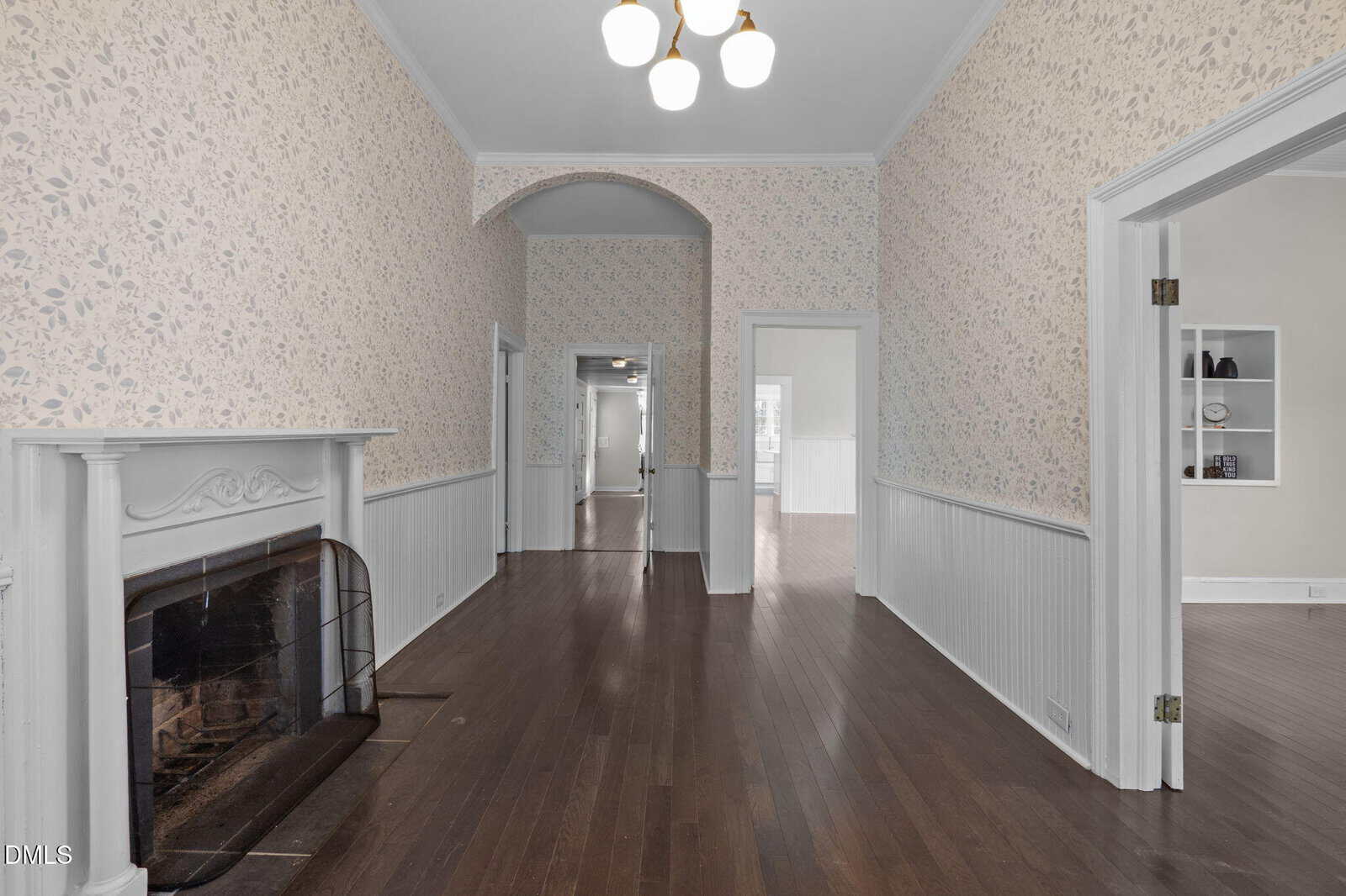 A hallway with dark wood floors, floral wallpaper, a white fireplace on the left, and a central archway. There are open doorways leading to adjacent rooms and ceiling lights overhead.