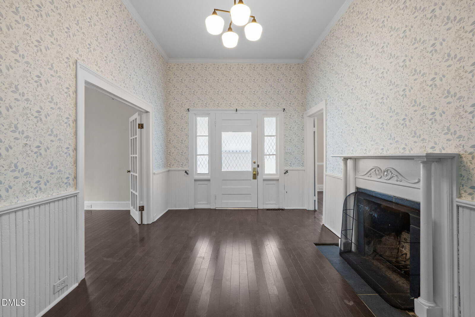 A bright room with dark wood floors, floral wallpaper, white paneling, a white front door with glass panes, a white fireplace, and a three-light ceiling fixture. Two open doorways lead to adjacent rooms.