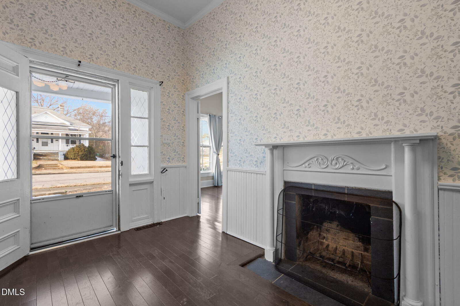 A bright room with floral wallpaper, a white fireplace, dark wood floors, and a glass-paneled door with sidelights that leads outside; another room with large windows is visible through an open doorway.