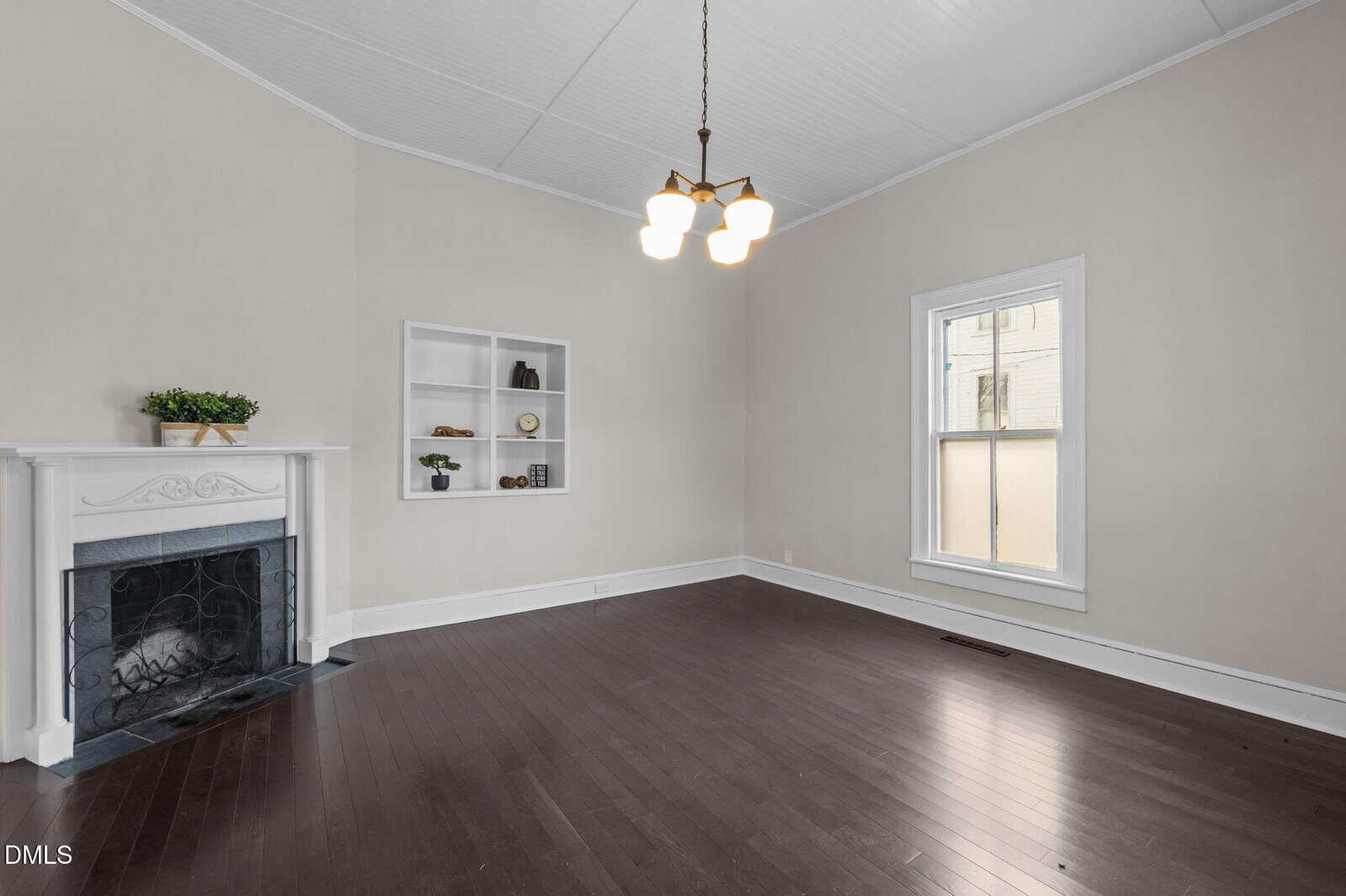 Spacious, empty living room with dark hardwood floors, light beige walls, tall white ceiling, a white fireplace with a potted plant, built-in shelves, a large window, and a central chandelier with four lights.