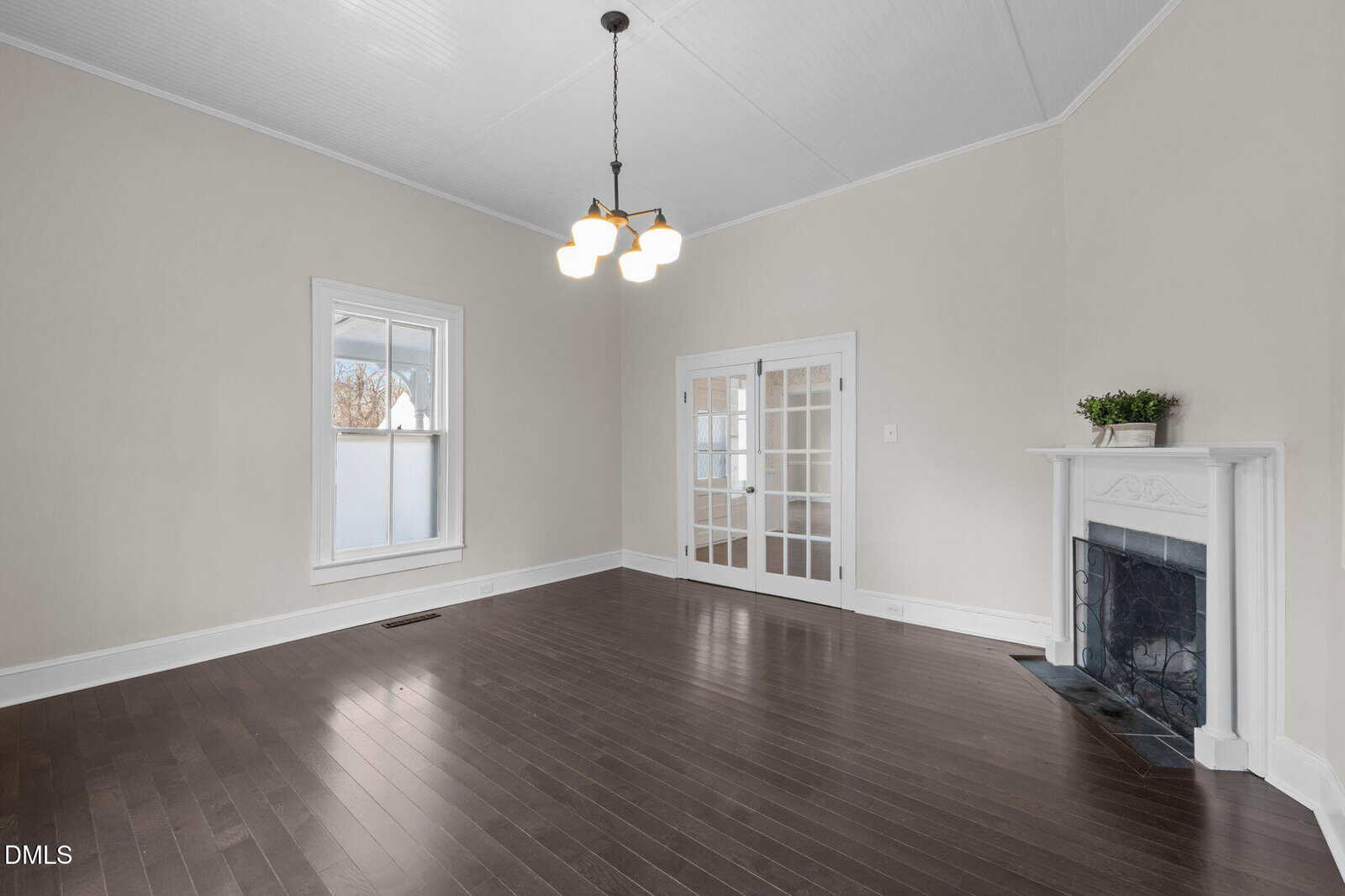 Bright empty room with dark hardwood floors, beige walls, white trim, a window, French doors, a white fireplace with a plant, and a ceiling light fixture.