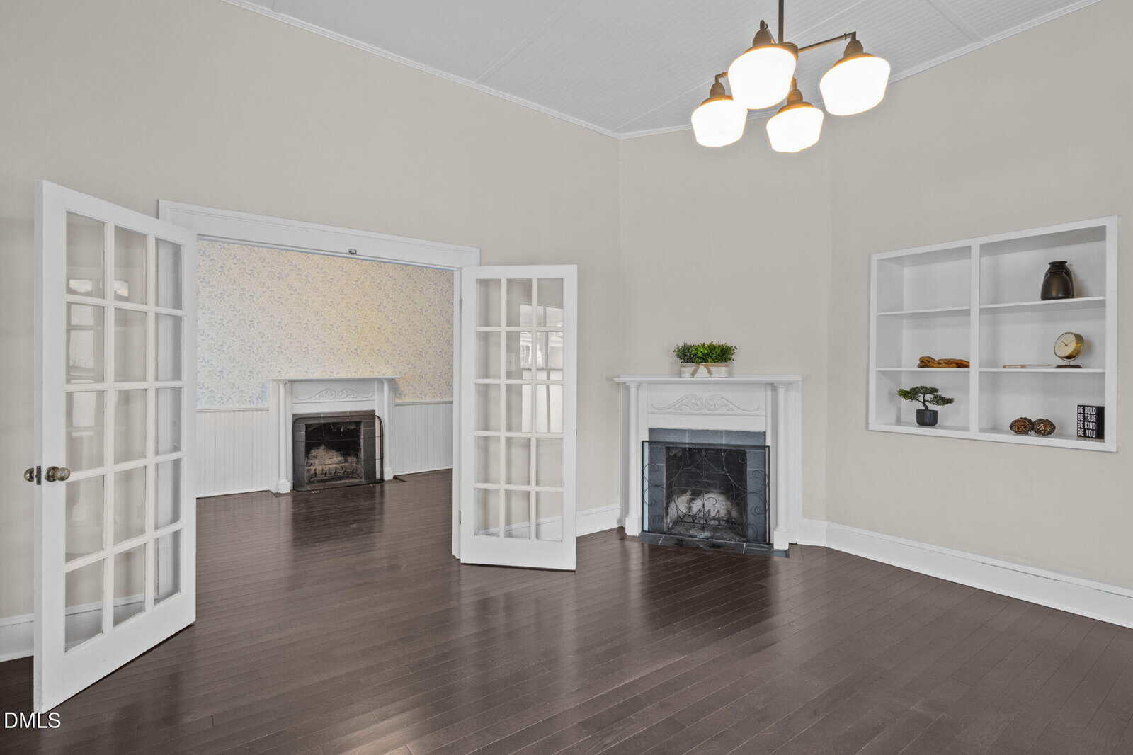 Bright living room with dark wood floors, white walls, French doors, two fireplaces, built-in shelves with decor, and a modern chandelier hanging from a high ceiling.