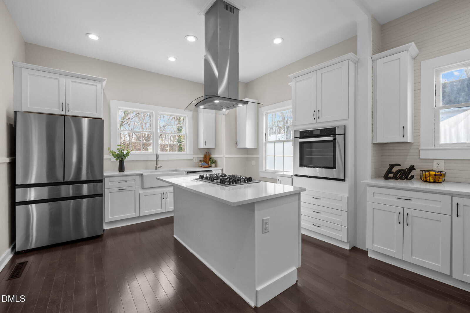 Modern kitchen with white cabinets, stainless steel appliances, a central island with a gas cooktop and hood, dark hardwood floors, and large windows letting in natural light.