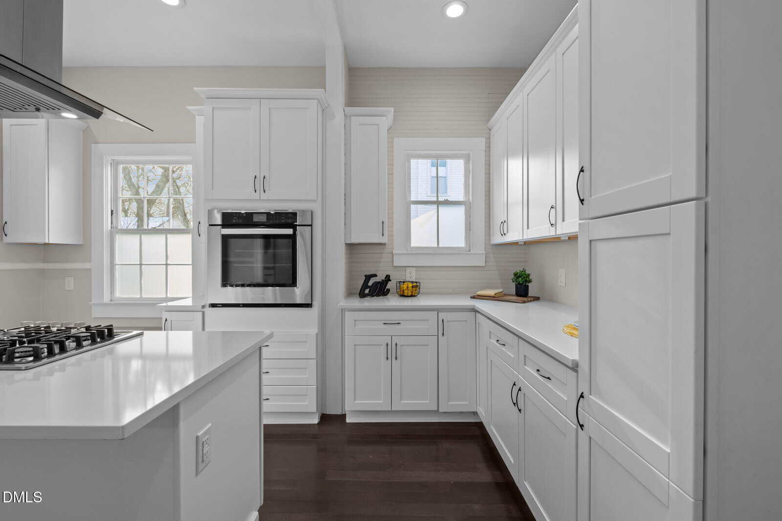 Modern kitchen with white cabinets, sleek built-in oven, gas stove on the island, dark wood floors, and two windows letting in natural light. Minimalist decor includes a bowl of lemons and some kitchen accessories.