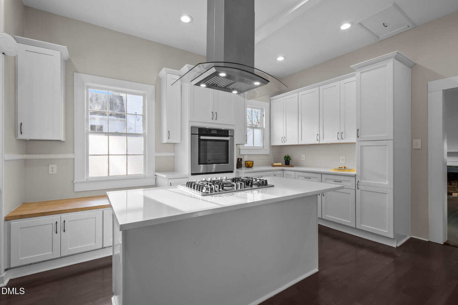 Modern kitchen with white cabinets, built-in wall oven, center island featuring a gas cooktop and range hood, white countertops, and large windows letting in natural light.