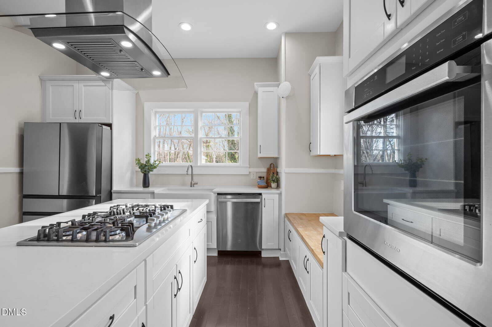 Modern kitchen with white cabinets, stainless steel appliances, a gas cooktop island, double wall oven, deep sink under two windows, and dark wood floors. A small plant sits on the windowsill.