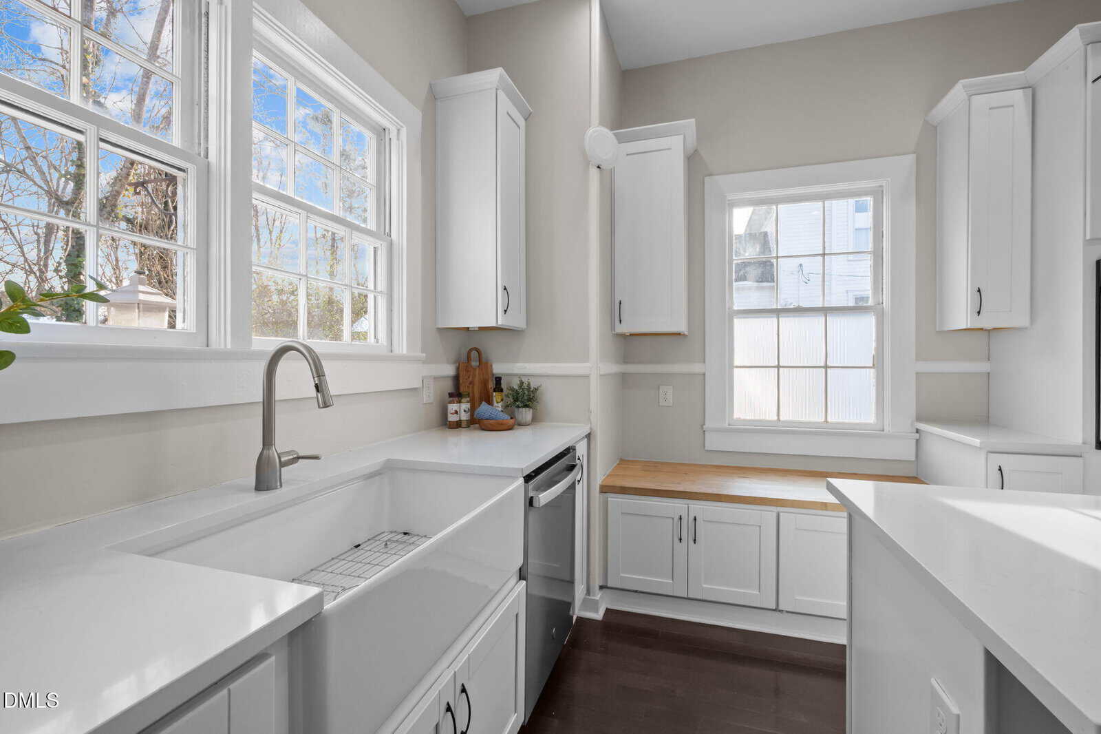 Bright, modern kitchen with white cabinets, white countertops, a farmhouse sink, stainless steel faucet, wood countertop along the window, and large windows letting in natural light.