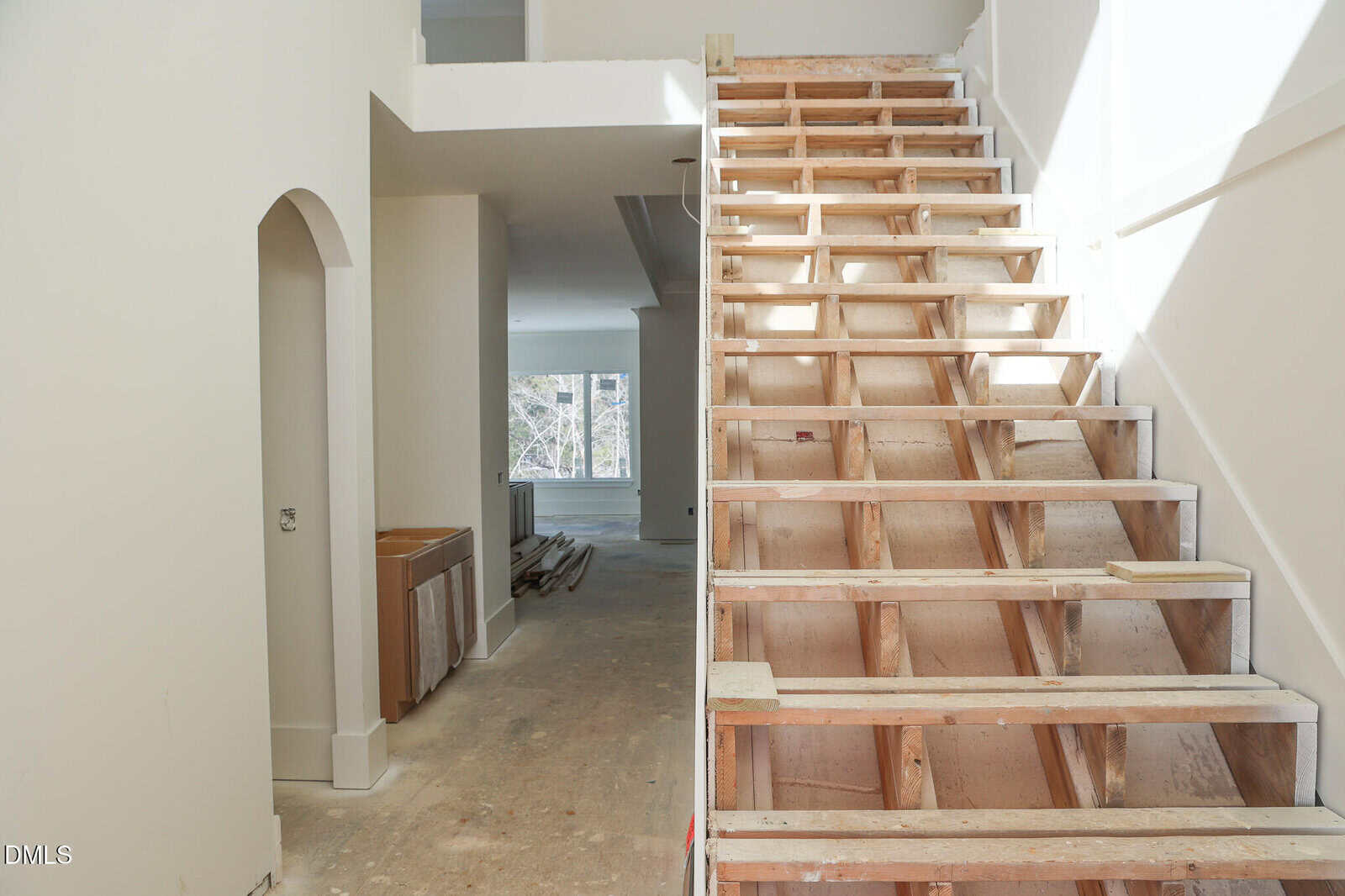 Unfinished staircase with exposed wooden framing in a house under construction. Sunlight streams through windows in the background, illuminating bare floors, unpainted walls, and installed cabinetry.
