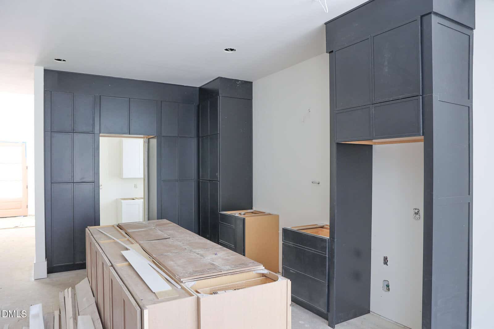 Modern kitchen under construction with unfinished cabinets, dark gray built-ins, and an island frame in the center. No appliances or countertops are installed yet. Walls are white and the floor is unfinished.