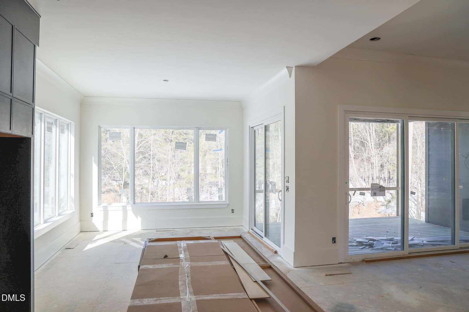 A bright, unfinished room with large windows and sliding glass doors, letting in natural light. The floor is bare with cardboard and wood planks scattered, and a partially completed deck is visible outside.