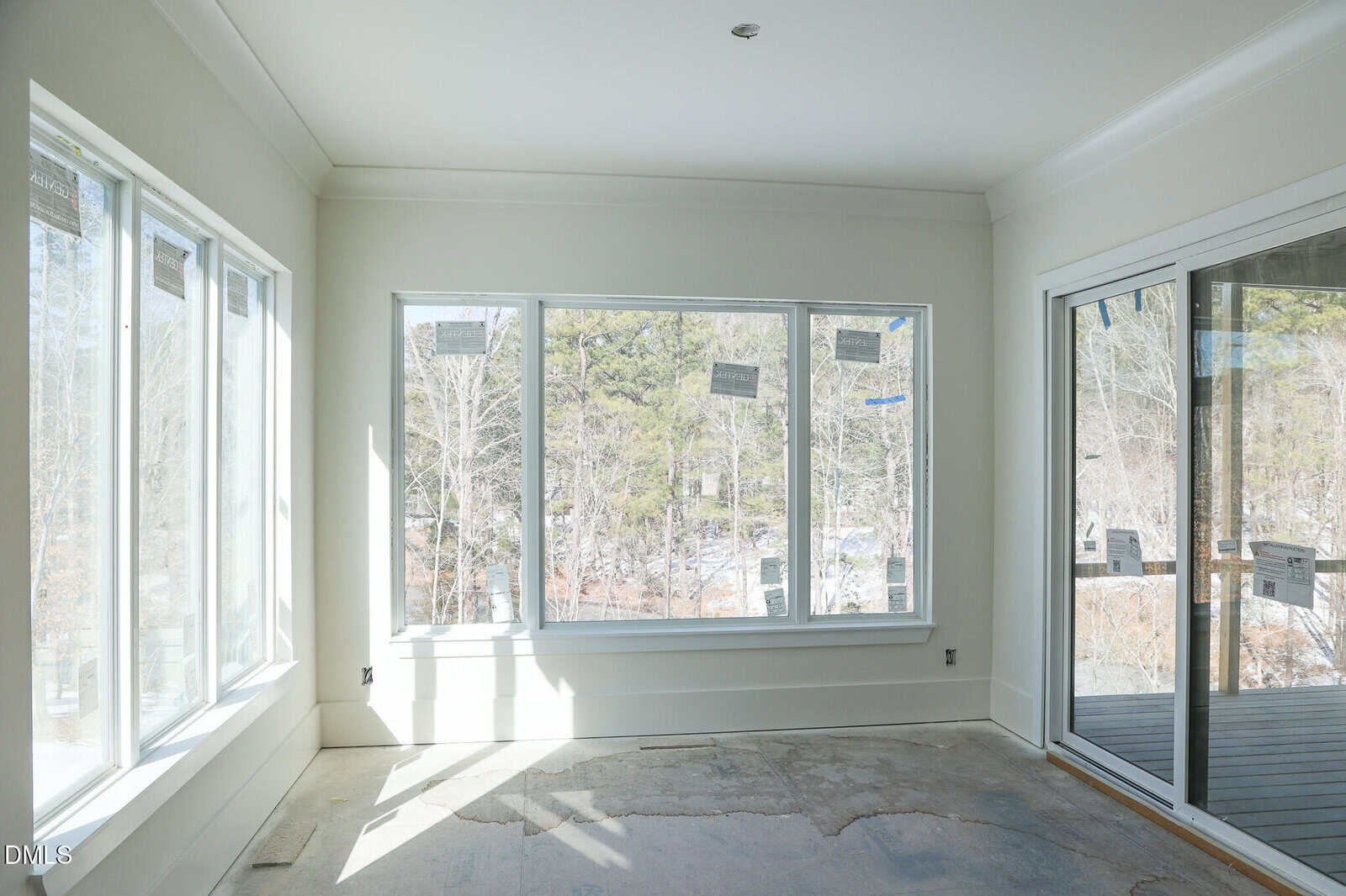 A bright sunroom with large windows, white walls, and an unfinished floor looks out onto a snowy, wooded landscape. Sliding glass doors open onto a deck.