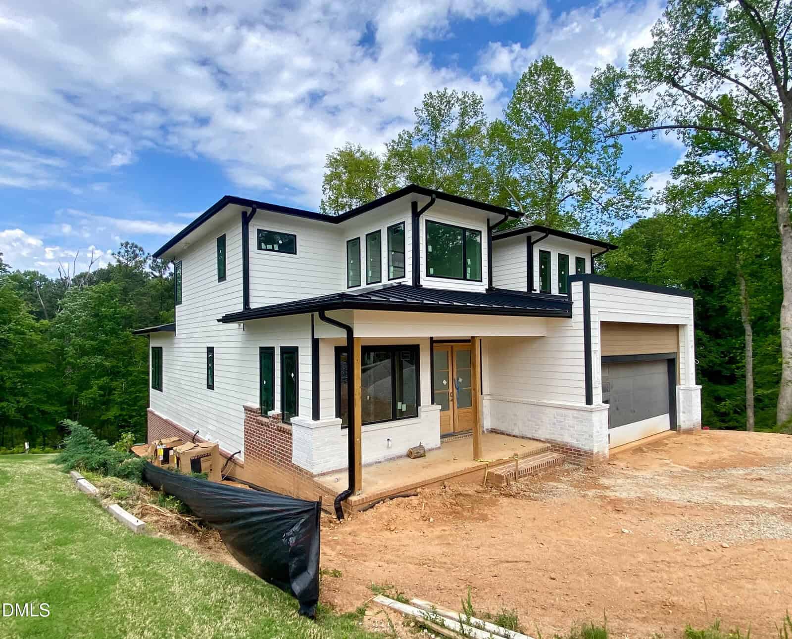 A modern two-story house with white siding, large windows, and a black roof sits on a dirt lot surrounded by trees, with a two-car garage and a wooden front door under a covered entry.