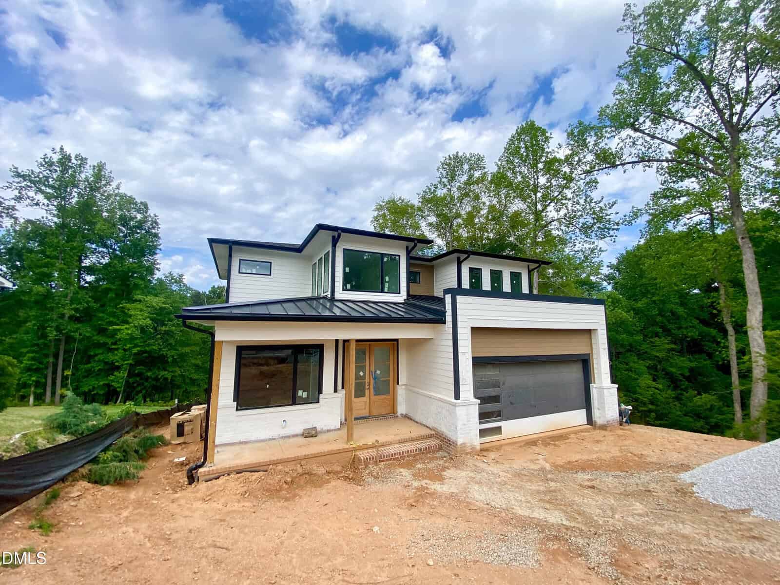 Modern two-story house with large windows, white siding, black trim and roof, a two-car garage, and wooden front doors. The house is surrounded by trees and an unfinished yard with dirt and gravel.
