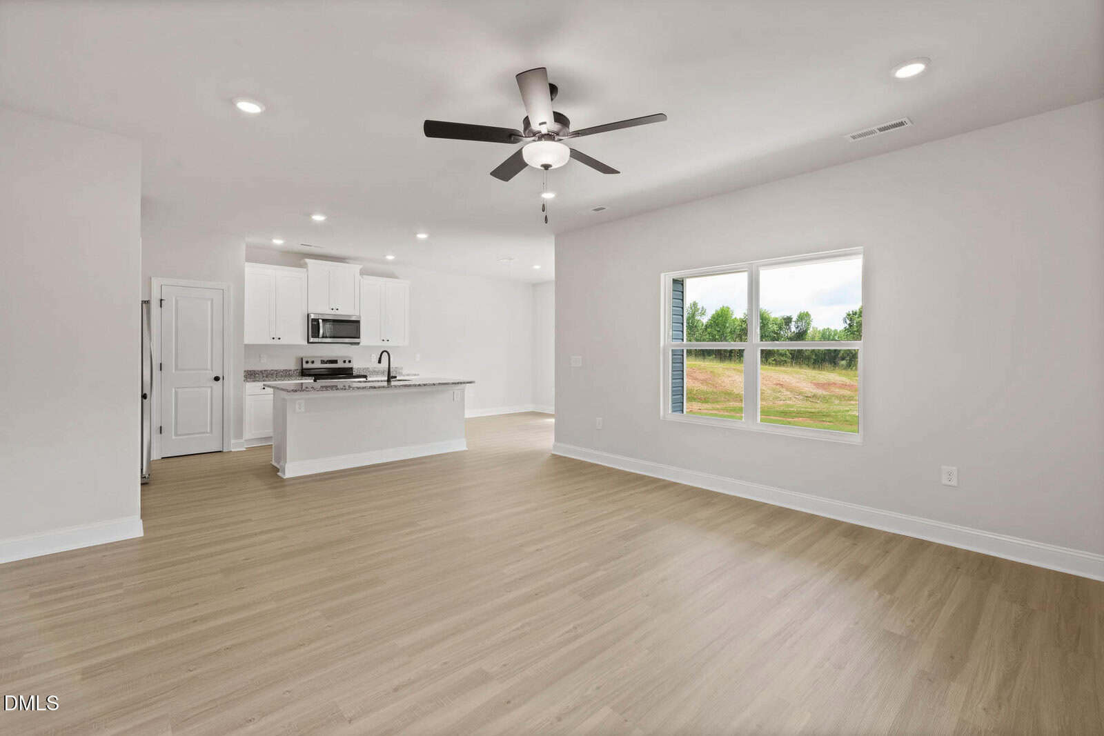 A spacious, unfurnished living area with light wood flooring, white walls, a ceiling fan, a large window, and an open kitchen with white cabinets and stainless steel appliances in the background.