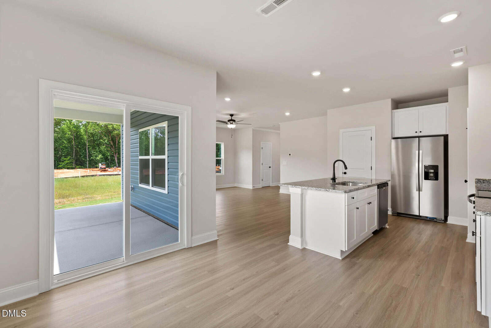 Open-concept kitchen with white cabinets, granite island, stainless steel appliances, and light wood floors; adjacent to a sliding glass door leading to a covered patio and backyard with trees visible outside.