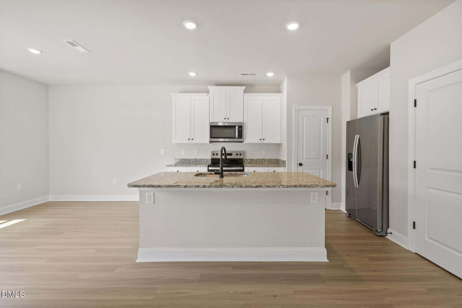 Modern kitchen with white cabinets, stainless steel appliances, granite countertops, an island with a sink, and light wood flooring. The space is well-lit with recessed ceiling lights.