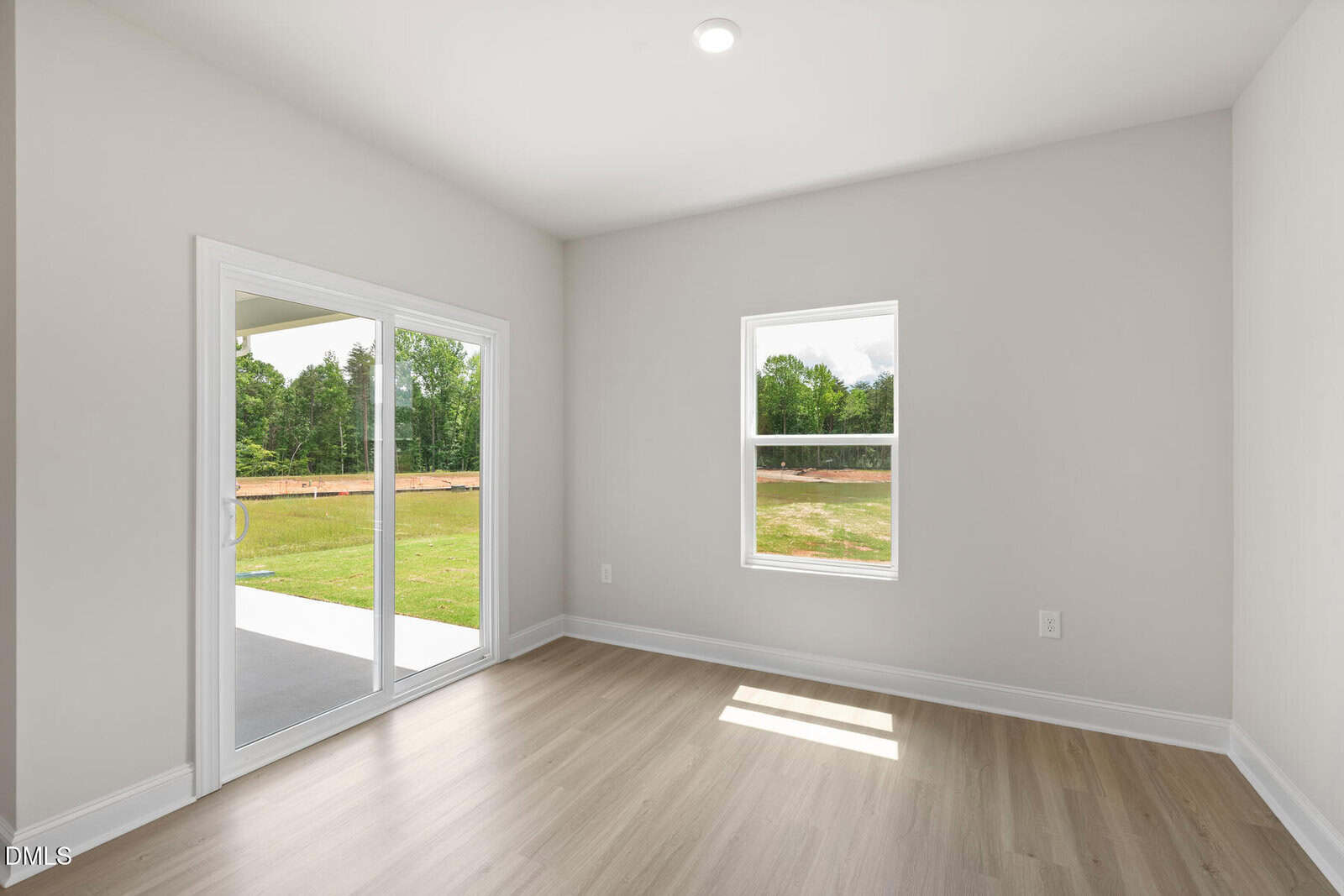 A bright, empty room with light wood flooring, white walls, a window, and sliding glass doors leading to a grassy backyard with trees. Natural light fills the space.