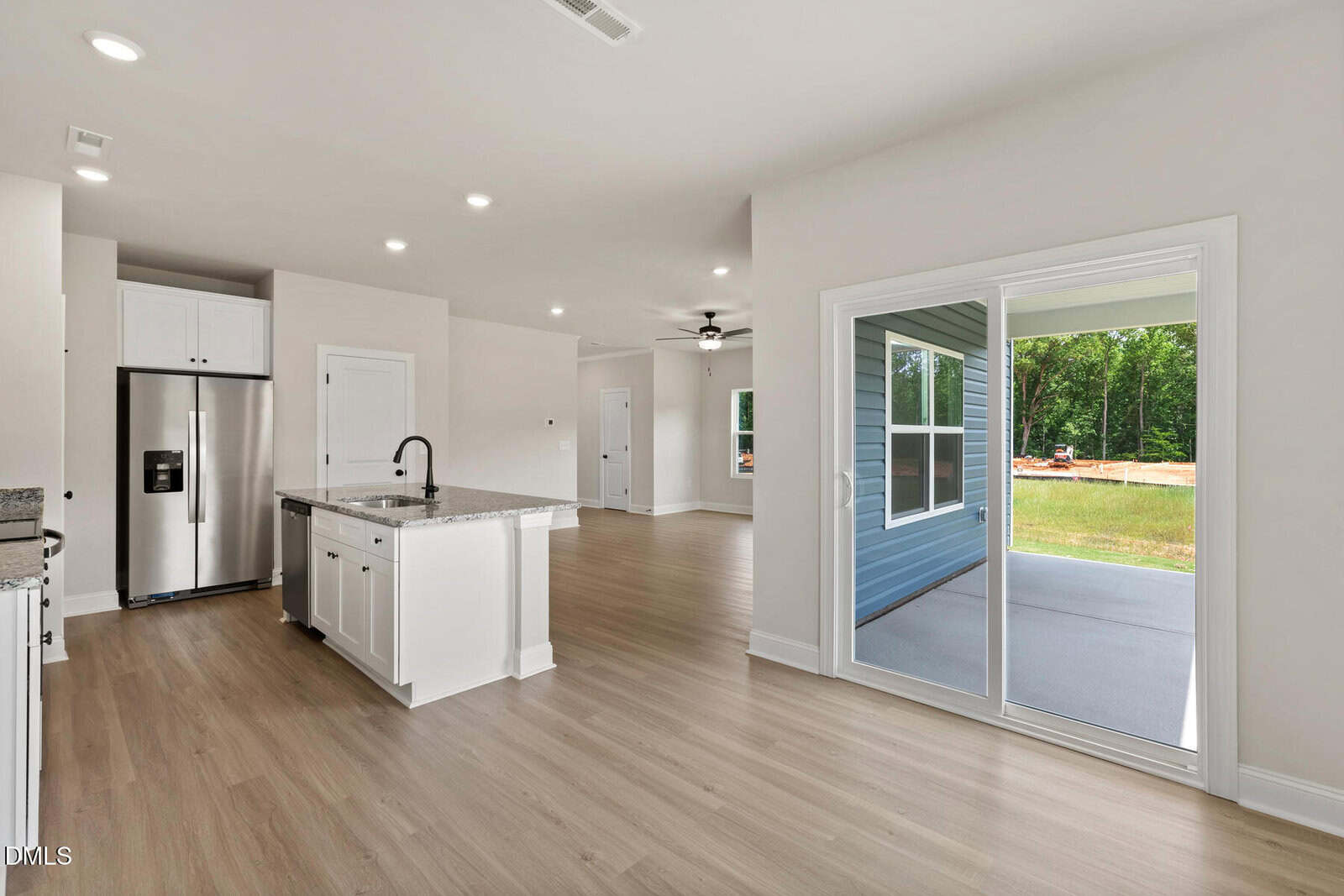 Modern, unfurnished kitchen and dining area with light wood floors, white cabinets, stainless steel appliances, and a center island. Sliding glass doors open to a covered patio and green outdoor space.