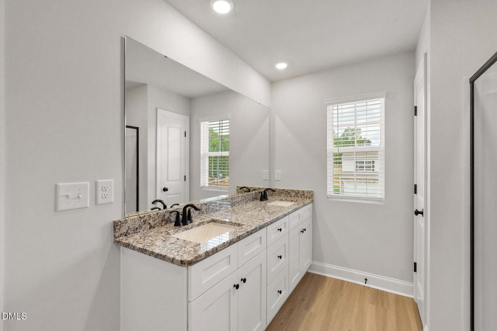 A modern bathroom with a granite double-sink vanity, white cabinets, black faucets, a large wall mirror, two windows with white blinds, and light wood flooring. The walls are painted light gray.