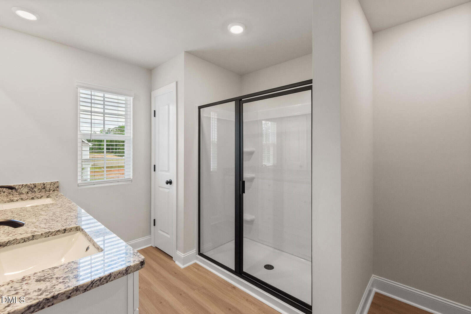 Modern bathroom with a glass-enclosed shower, double sink with granite countertop, white cabinets, wood flooring, a window with blinds, and white walls.