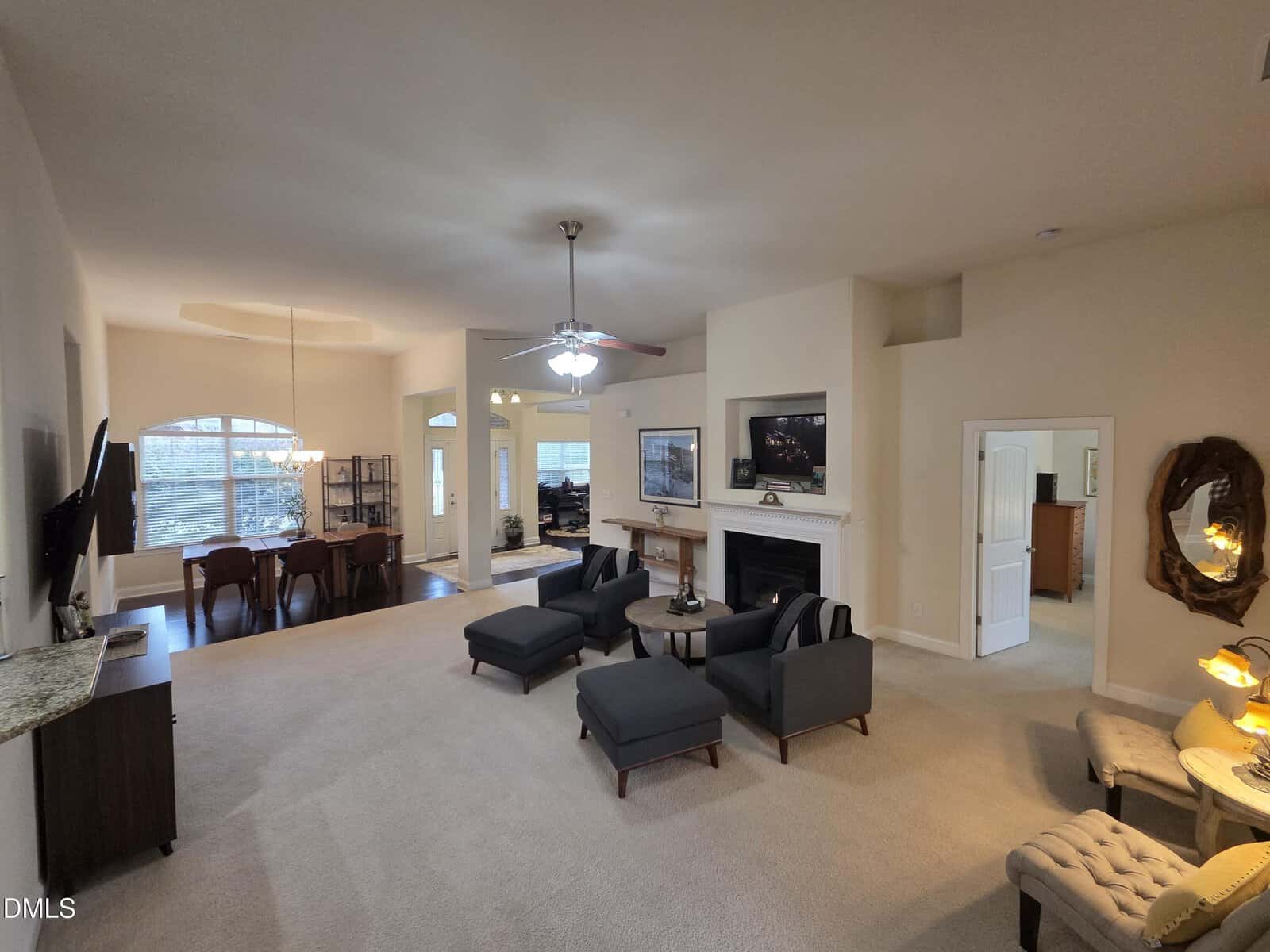 Spacious open-concept living area with beige carpet, modern gray furniture, ceiling fan, fireplace with TV above, dining area to the left, and multiple rooms visible through open doorways. Large windows bring in natural light.