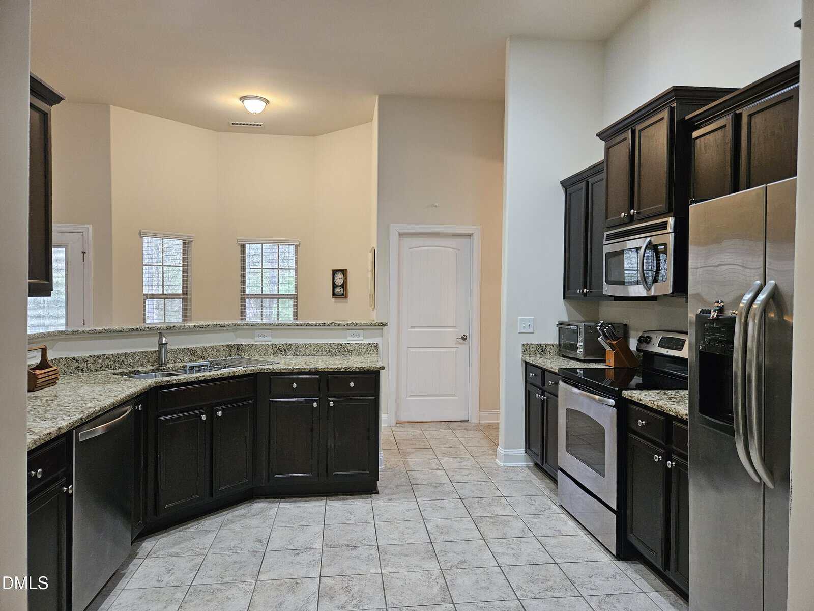 Modern kitchen with dark cabinets, granite countertops, stainless steel appliances, and a tiled floor. The room features a double sink, microwave, oven, refrigerator, and three large windows providing natural light.