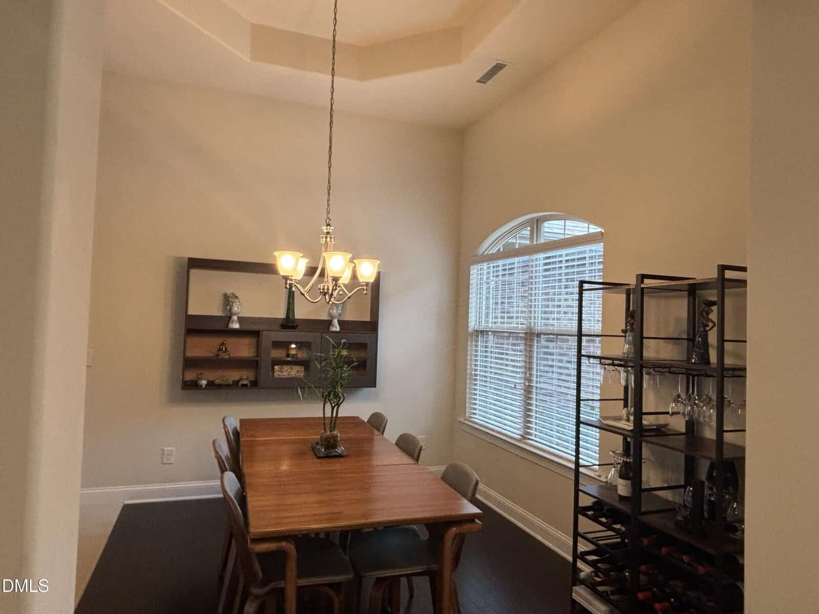 Dining room with a wooden table and six chairs, a chandelier overhead, a large window with blinds, a shelving unit holding wine bottles and glasses, and a wall-mounted display shelf with decorative items.