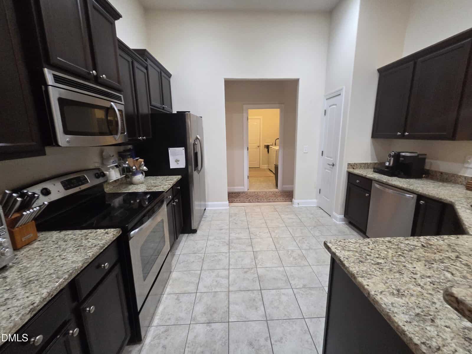 A modern kitchen with dark wood cabinets, stainless steel appliances, granite countertops, and beige tiled floors. The kitchen opens to a hallway and another room in the background.