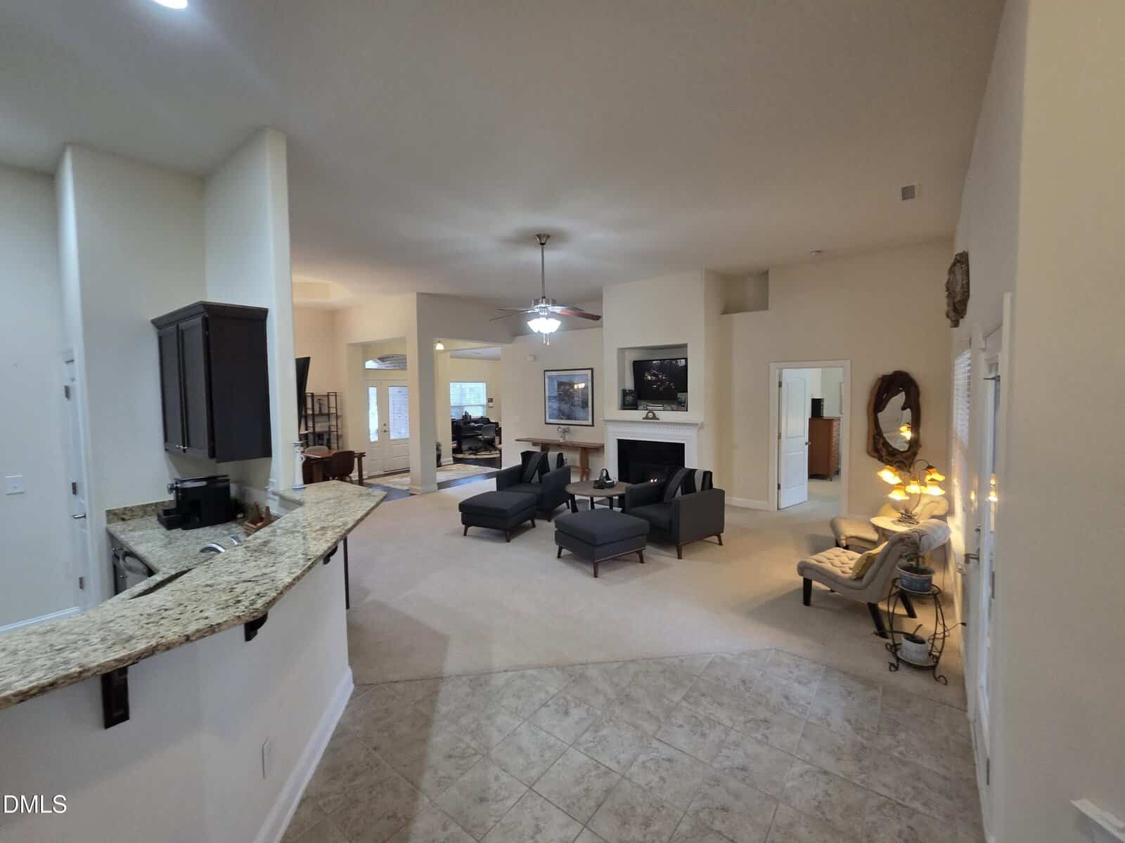 Open-concept living area with a granite kitchen counter on the left, tile and carpet flooring, cozy seating around a fireplace, ceiling fan, and multiple doorways leading to other rooms. Warm lighting and neutral decor throughout.