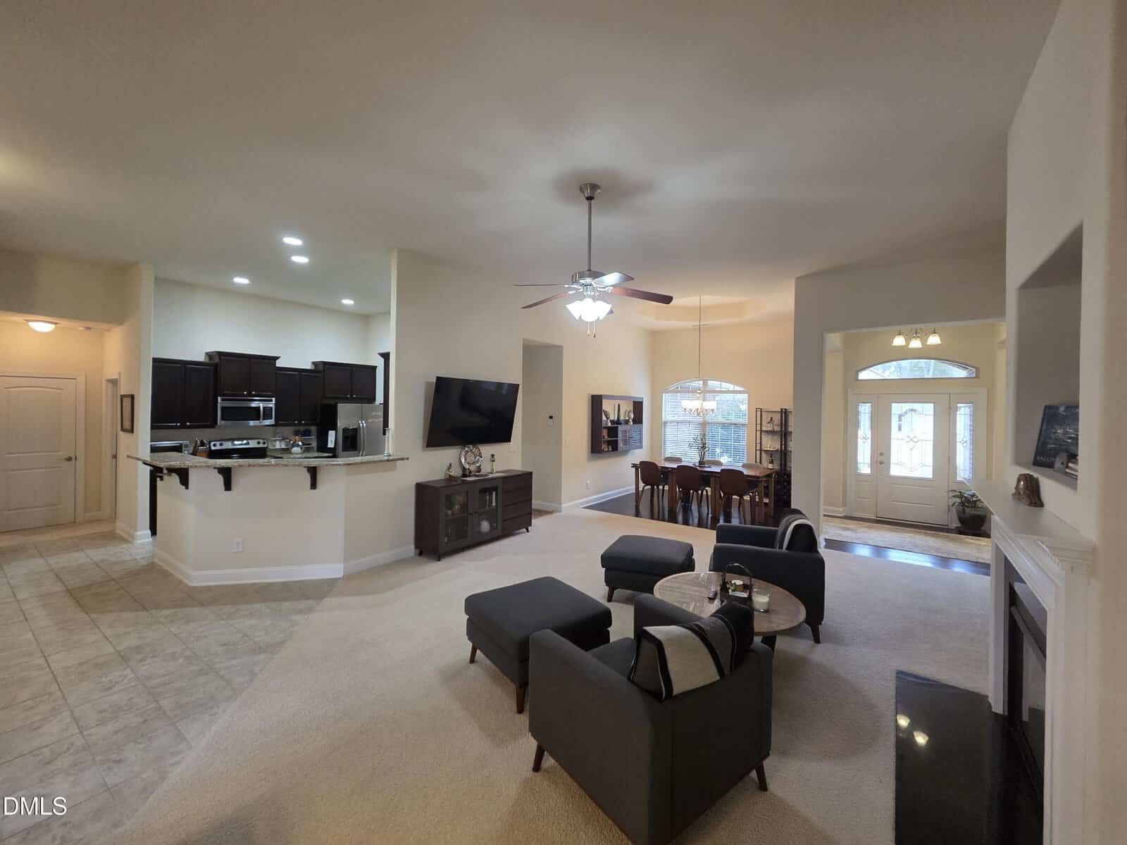 Open-concept living area with a kitchen, dining space, and sitting area. Dark cabinets, granite counters, and bar stools in the kitchen. Living room has modern seating, a TV, and ceiling fan. Natural light from large windows and a glass front door.