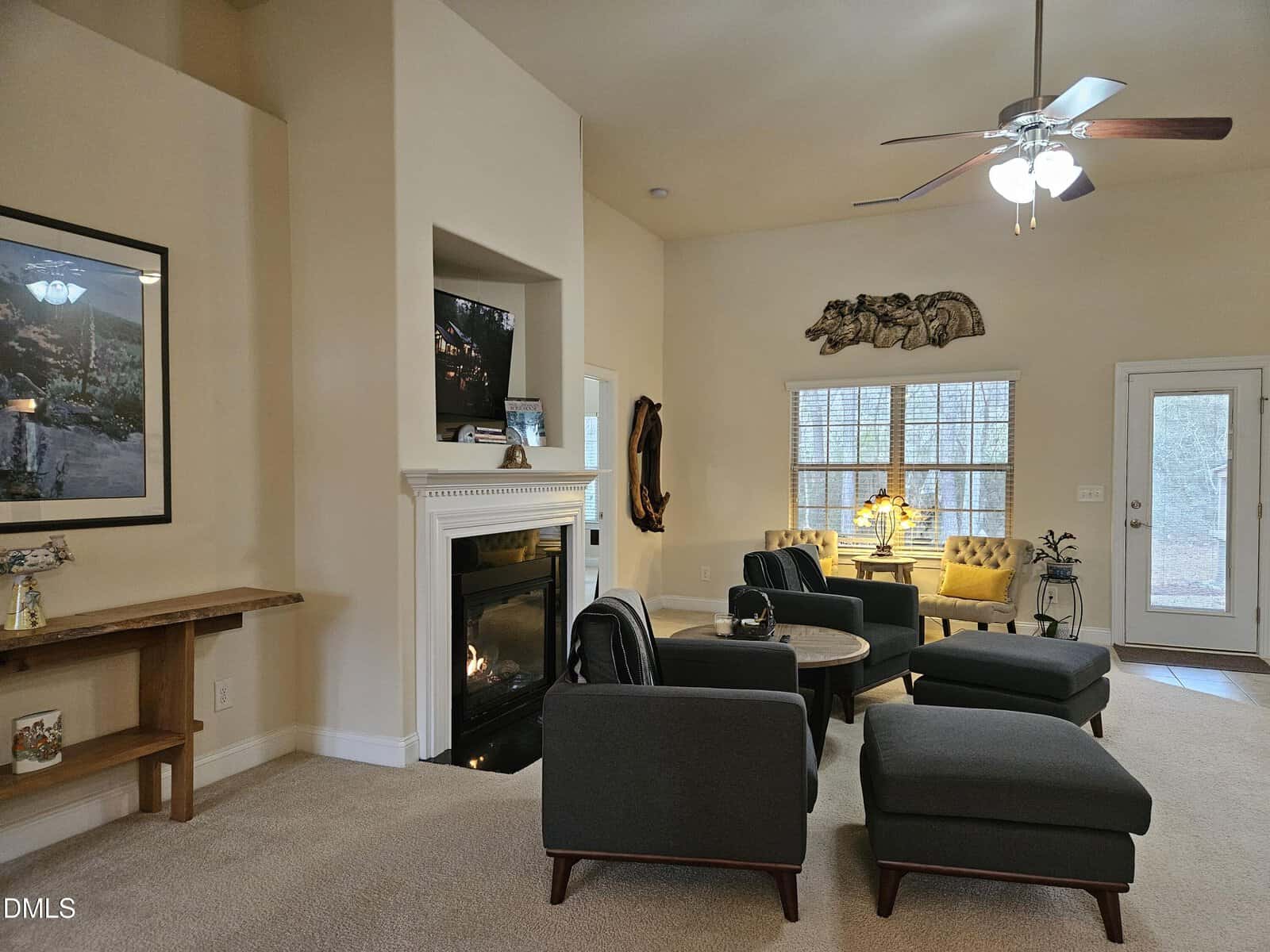 A cozy living room with a fireplace, dark modern chairs, a round coffee table, and ottomans. Large windows let in natural light. A ceiling fan hangs overhead, and art decorates the light-colored walls.