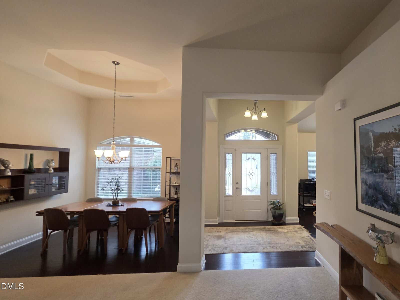 Spacious entryway and dining room with large windows, a wooden dining table with six chairs, chandelier, shelves, a white front door with glass panels, dark wood floors, and light-colored walls.
