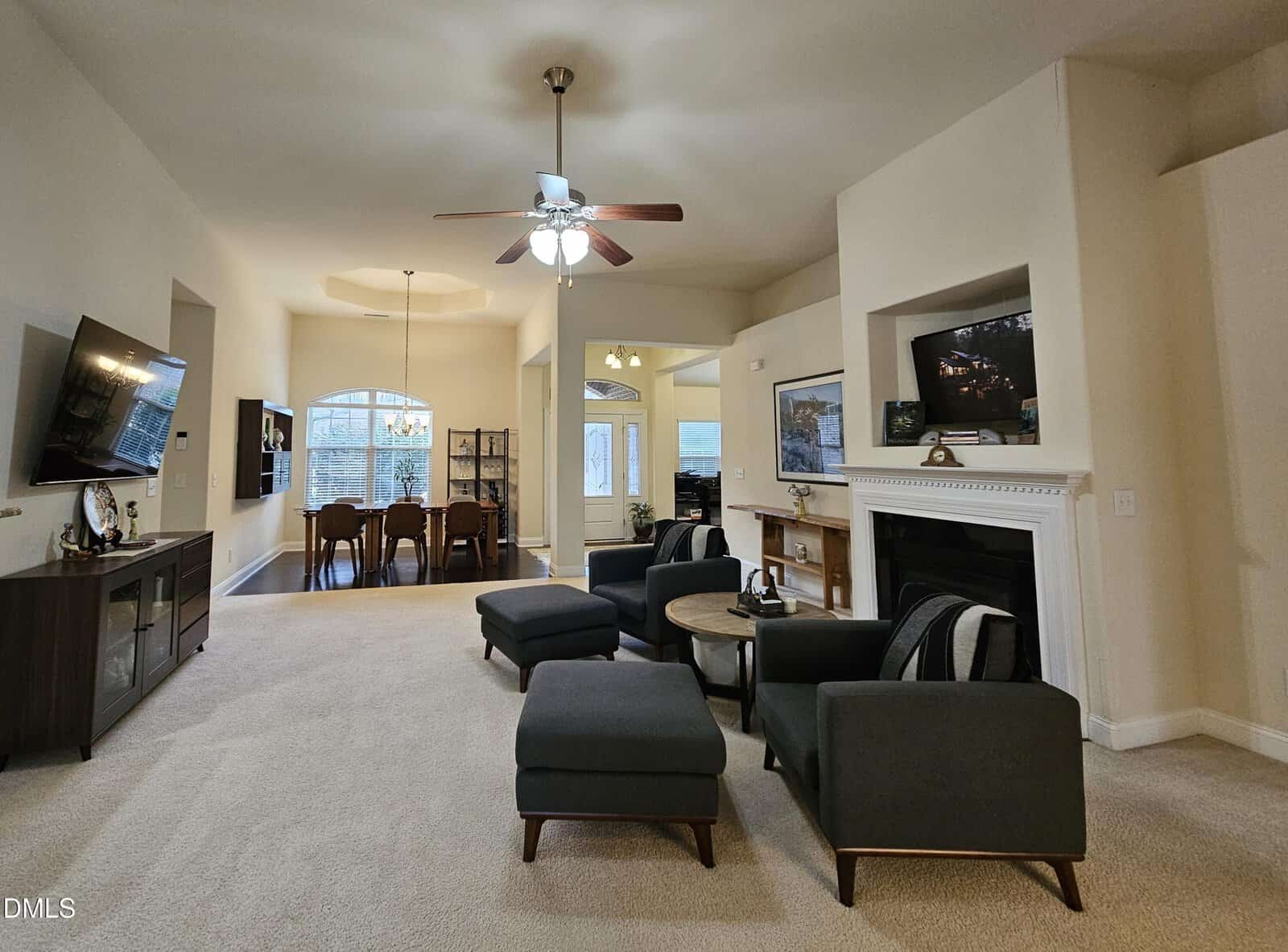 Spacious living room with dark armchairs and ottomans, a fireplace with a TV above, and a ceiling fan. Dining area with a large window is visible in the background. Neutral walls and carpet throughout.
