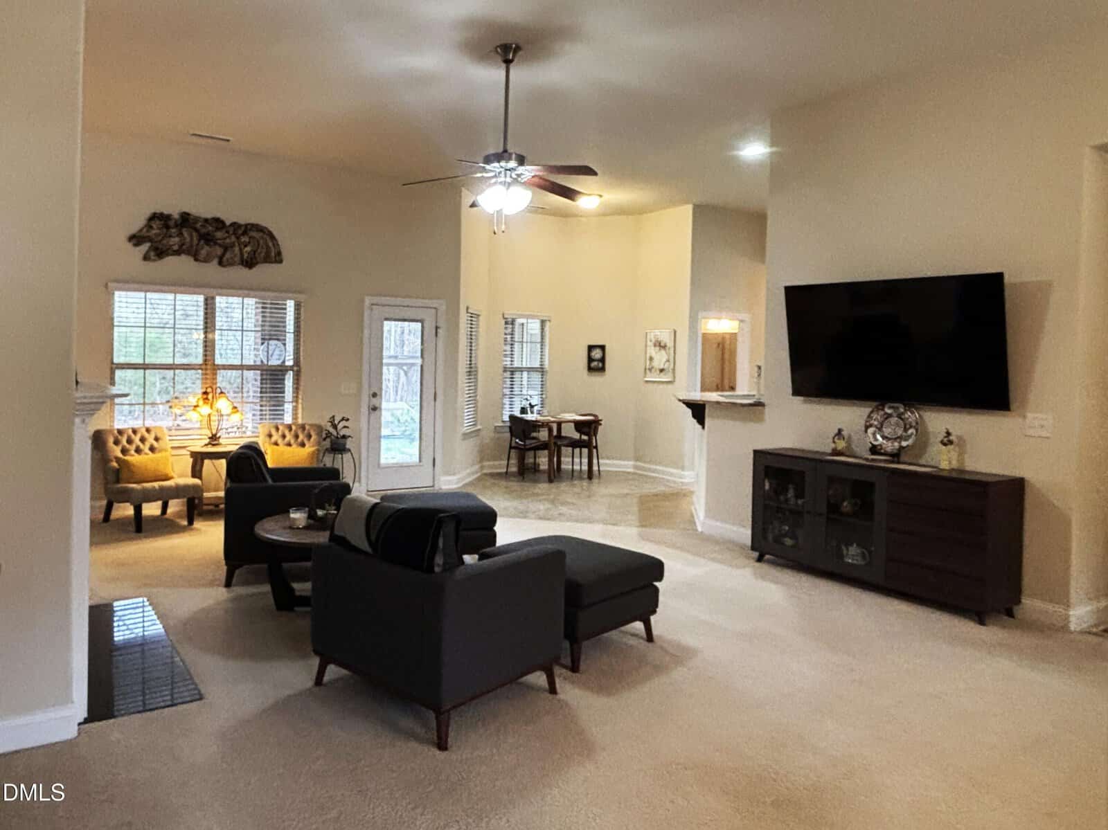 A spacious living room with beige walls and carpet, featuring a gray sectional sofa, armchairs, a wall-mounted TV, and large windows providing natural light. A dining area is visible in the background near glass doors.