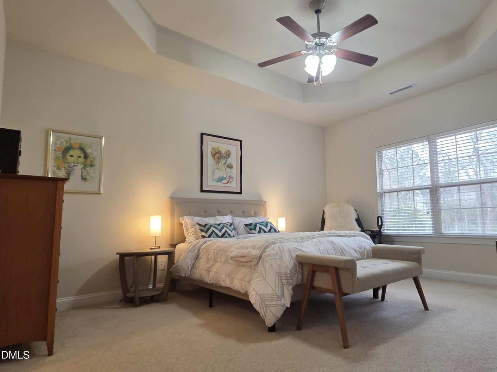 A neatly made bed with patterned pillows sits in a spacious, carpeted bedroom with beige walls, artwork, a ceiling fan, and large windows letting in natural light.