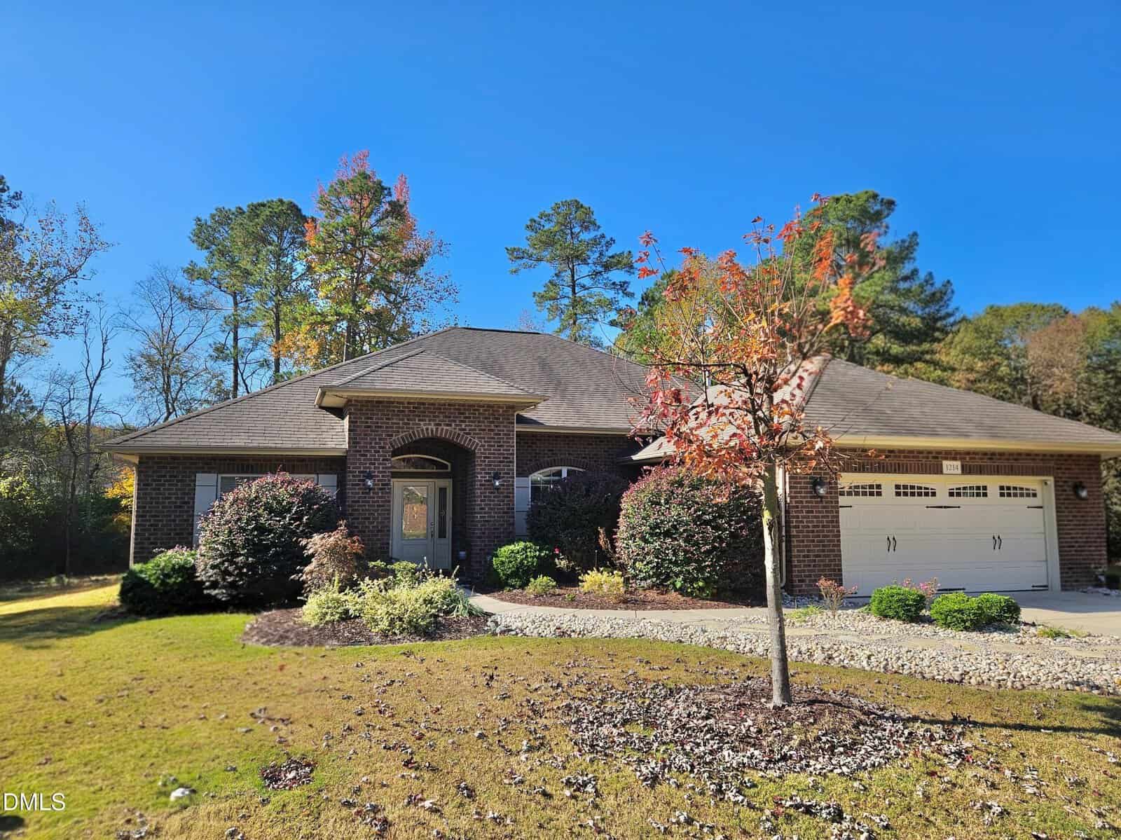 A single-story brick house with a two-car garage, manicured shrubs, a small tree, and a stone-bordered lawn under a clear blue sky with tall trees in the background.