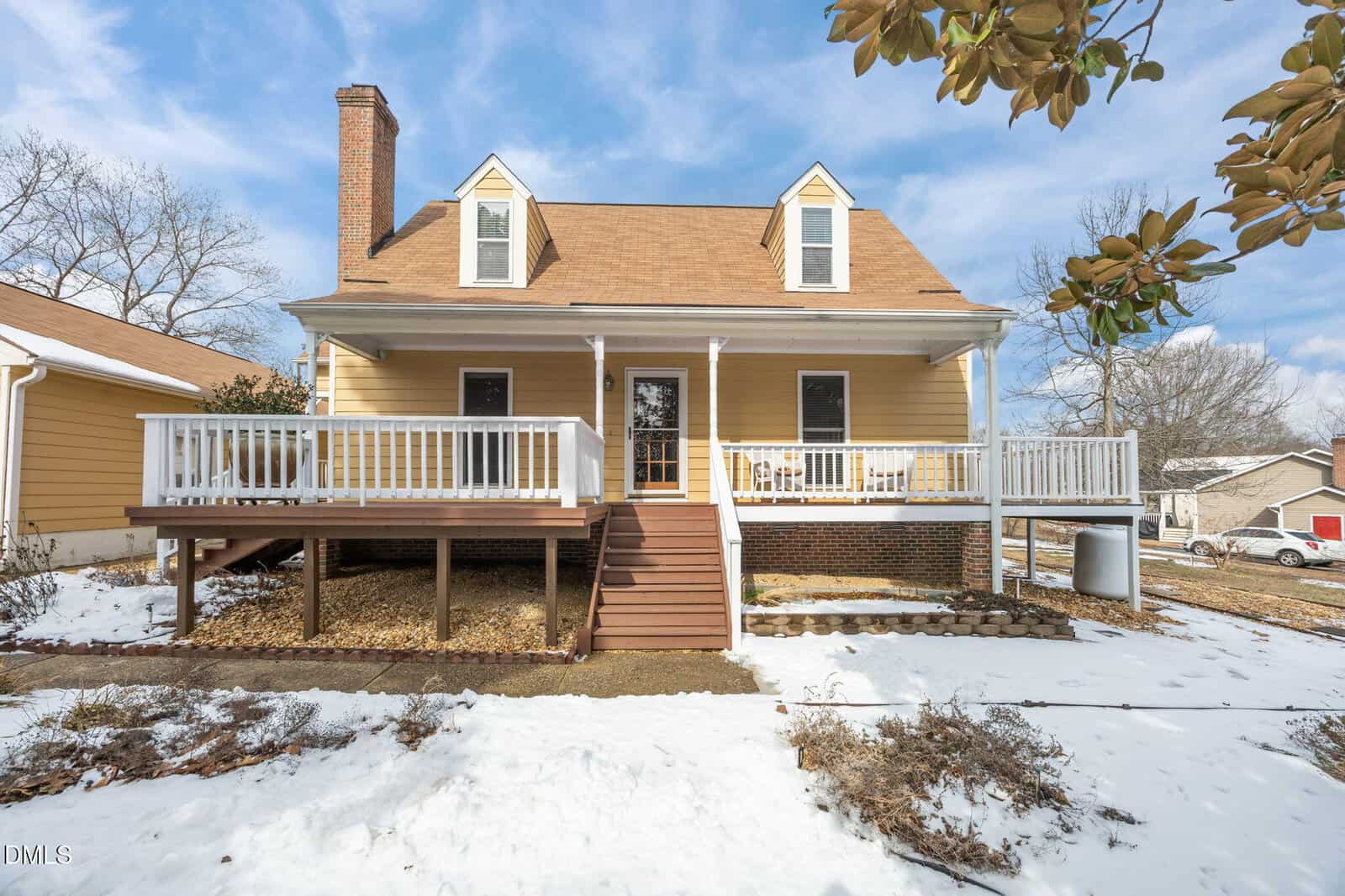 A yellow two-story house with a brown roof, white trim, and a large front porch sits on a snowy lawn. Steps lead up to the porch, and there are bare trees and a clear blue sky in the background.