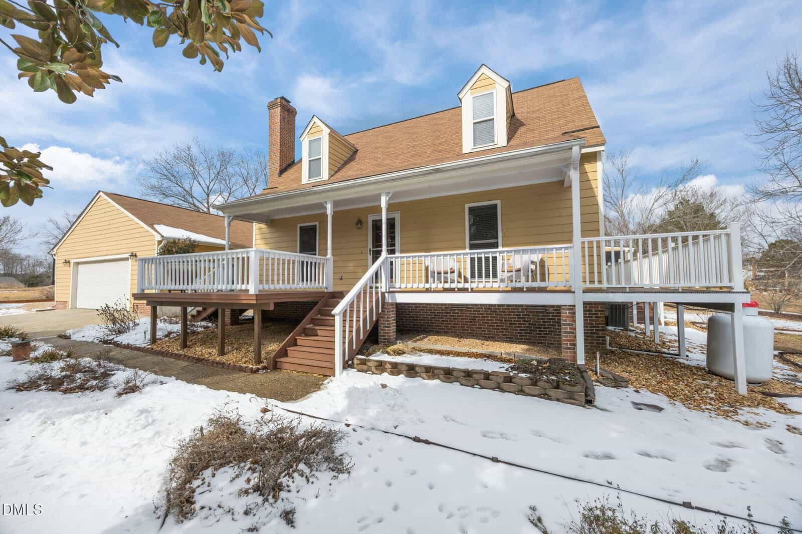 A two-story house with a large front porch, white railings, tan siding, and a brown roof. Snow covers the ground, and a detached garage is visible to the left. Leafless trees and a blue sky are in the background.