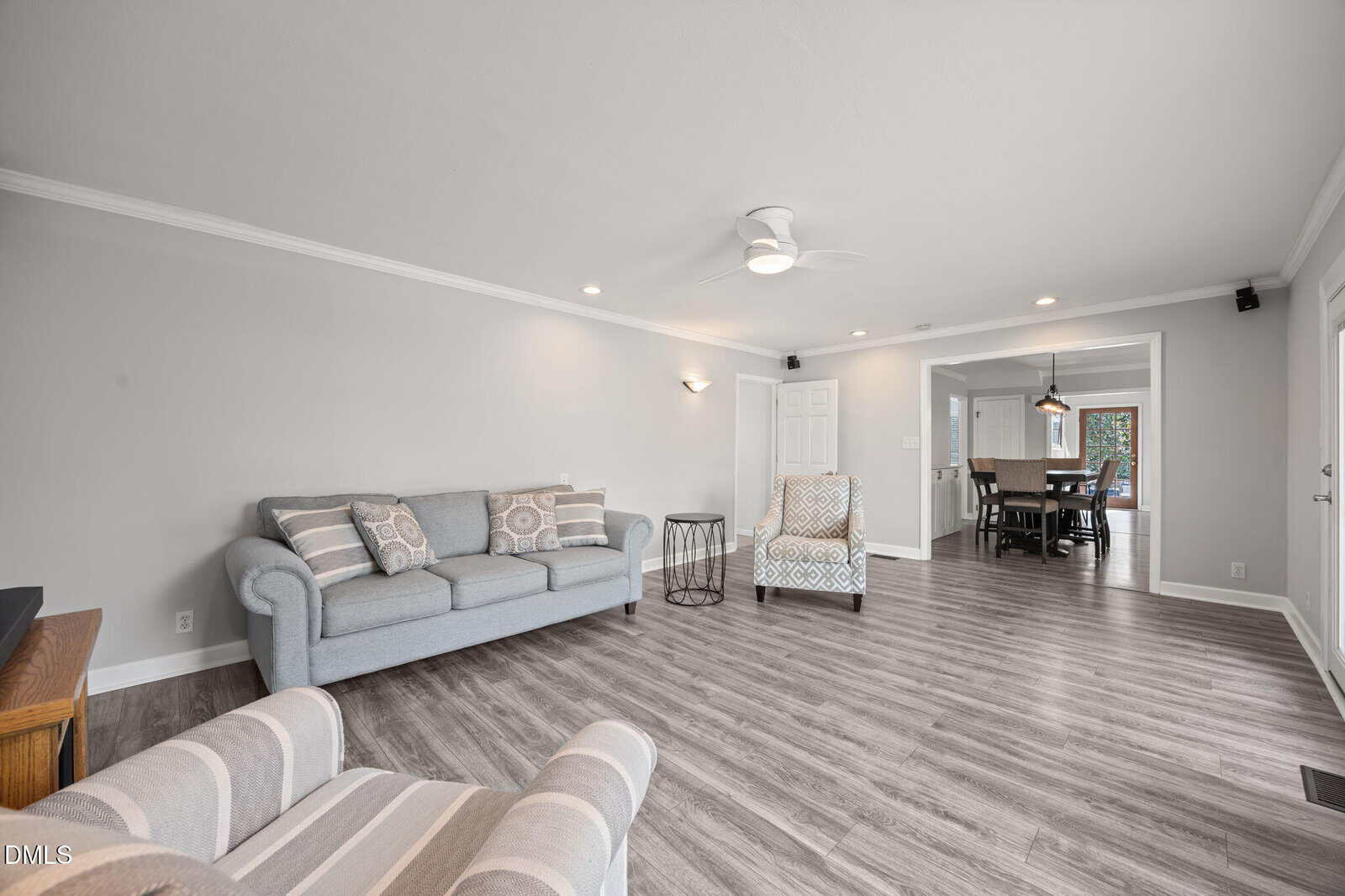 A bright living room with gray walls, wood-style flooring, a light gray sofa, two patterned armchairs, a round side table, and recessed ceiling lights. The room opens to a dining area in the background.