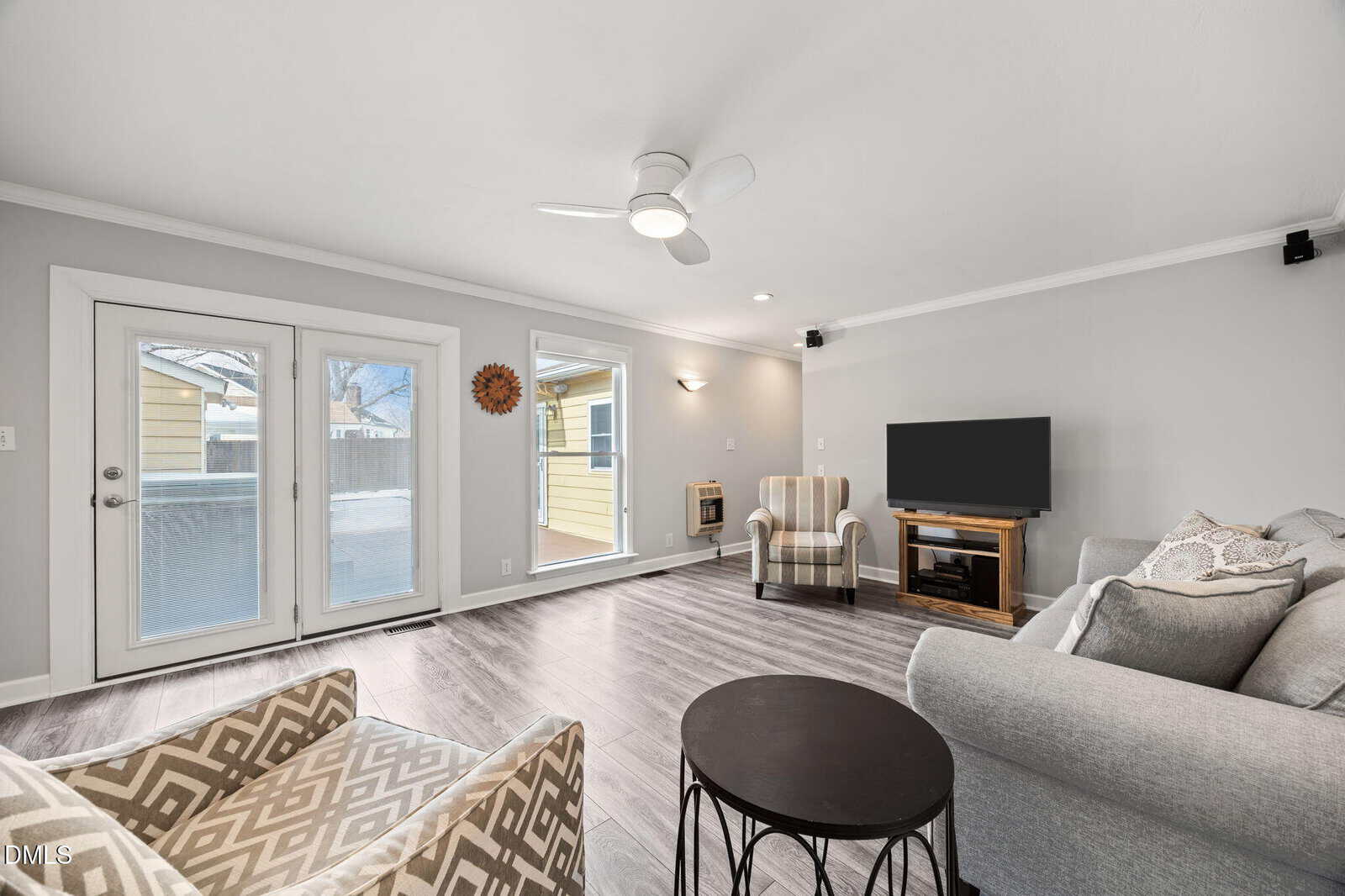 Bright living room with gray walls, wood flooring, a gray sofa, patterned armchair, black round coffee table, TV on a wooden stand, French doors, and large windows letting in natural light.