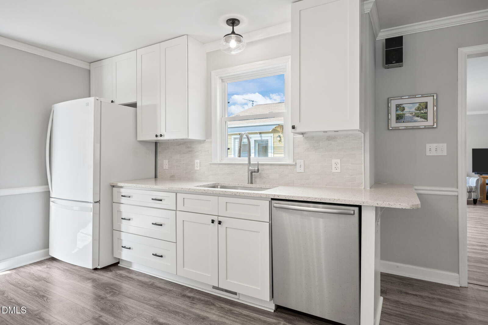 Modern kitchen with white cabinets, white refrigerator, stainless steel dishwasher, large window above sink, and light gray walls. Bright natural light shines through the window, and wood-like flooring is visible.