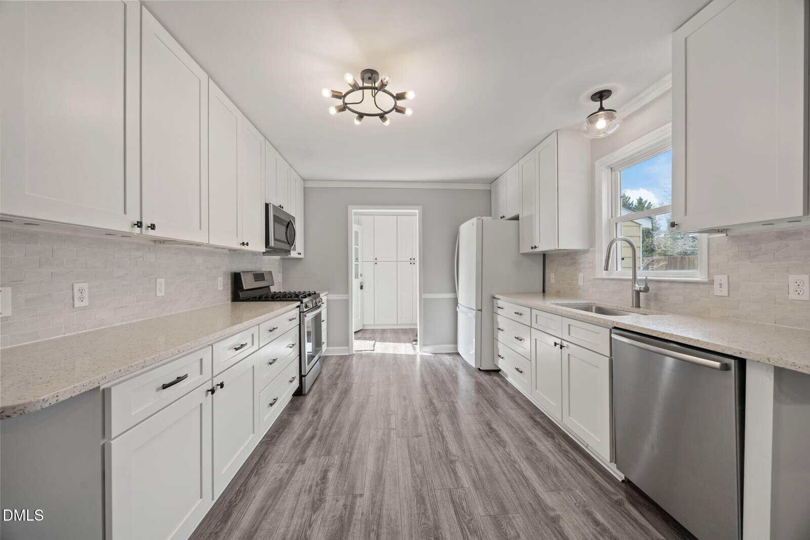 Modern kitchen with white cabinets, stainless steel appliances, light quartz countertops, a tile backsplash, large window over the sink, wood-look flooring, and a central ceiling light fixture. Doorway leads to another room.