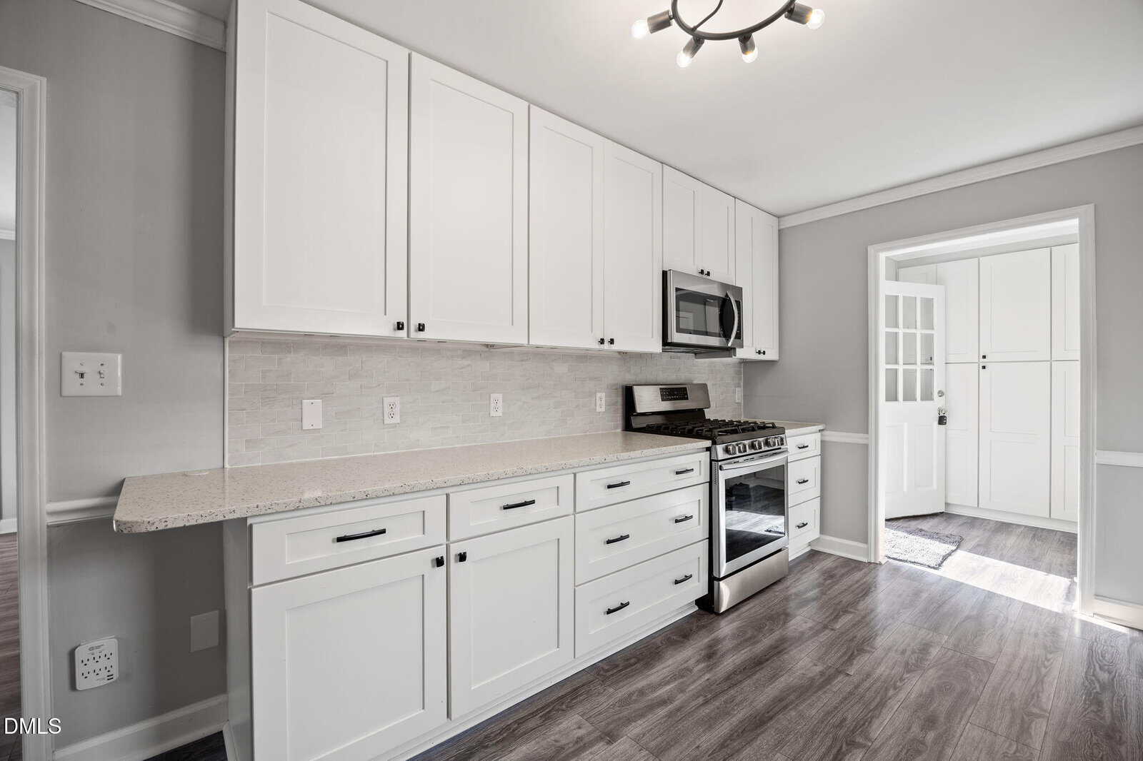 Modern kitchen with white cabinets, stainless steel stove and microwave, light countertops, tile backsplash, and wood flooring. A door and entryway are visible in the background.