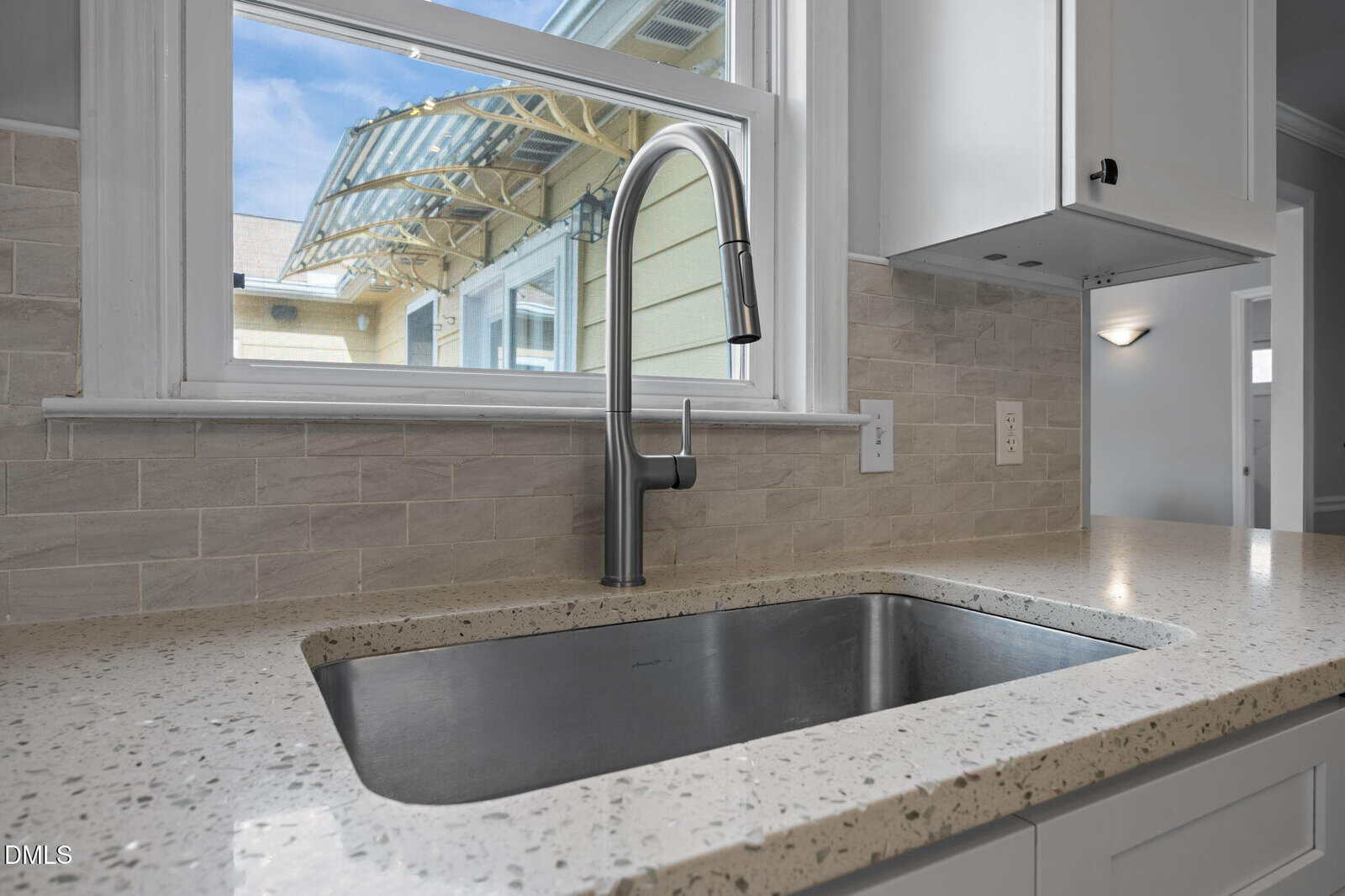 Modern kitchen sink with a sleek stainless steel faucet set in a light speckled countertop, in front of a window showing part of an exterior roof and blue sky. White cabinets and tile backsplash are also visible.