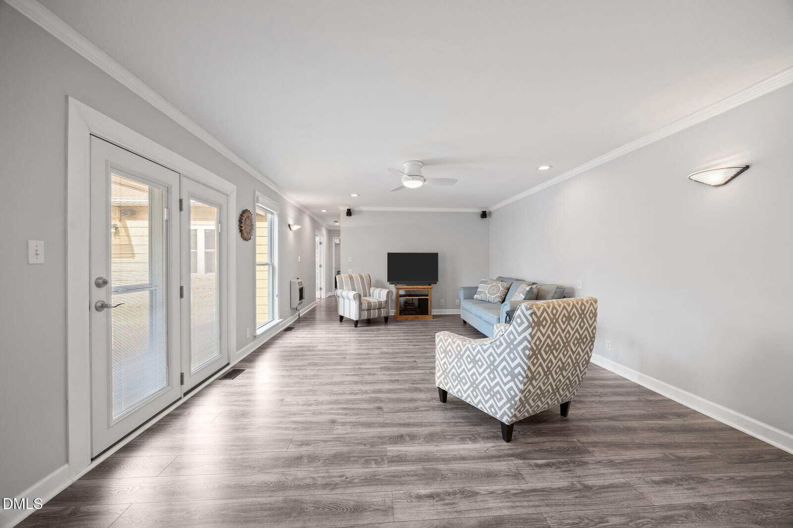 A spacious living room with light gray walls, wood flooring, large windows, and glass doors. There are two patterned armchairs, a blue sofa, a TV on a stand, and modern ceiling and wall lighting.
