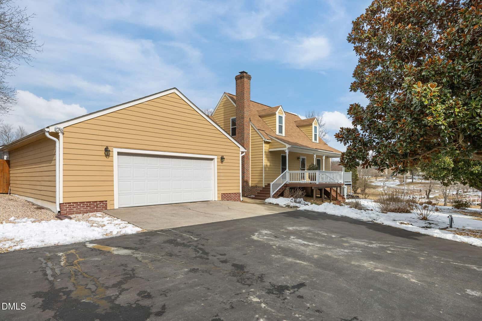 Yellow two-story house with a white garage, brick chimney, front porch, and snowy yard. Large tree on the right, paved driveway in front, and patches of snow on the ground under a partly cloudy sky.