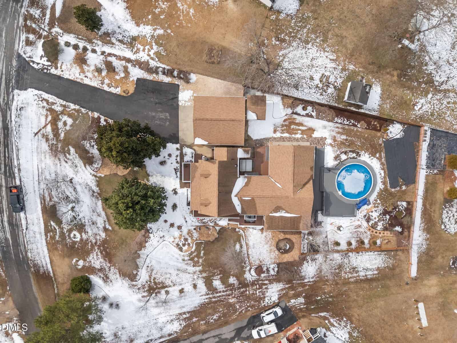 Aerial view of a house with a tan roof, patches of snow covering the yard, a round above-ground pool, surrounding trees, a driveway, and several parked vehicles.