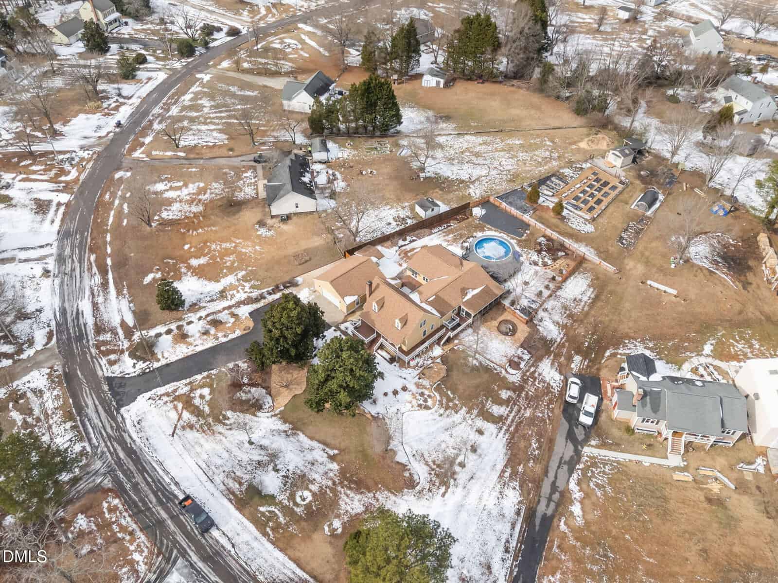 Aerial view of a suburban neighborhood in winter, with houses, yards, scattered snow patches, a round above-ground pool, gardens, and bare trees, all connected by winding roads.