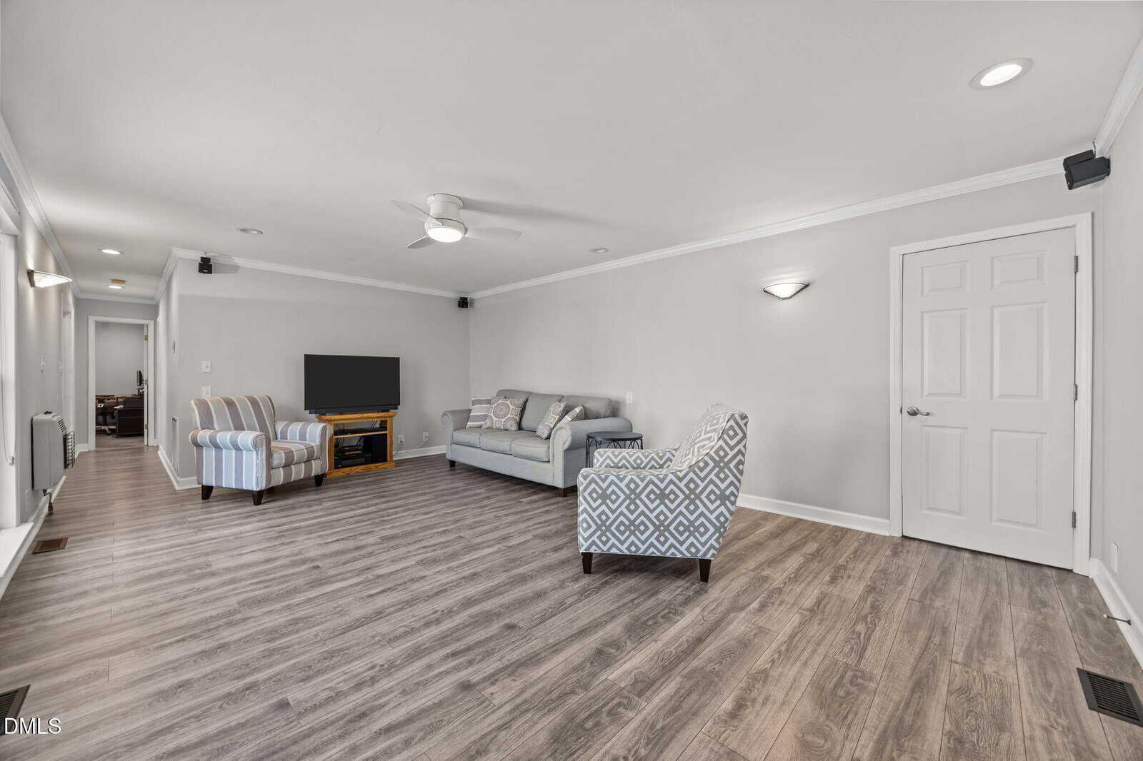 A spacious living room with light gray walls, wood-style flooring, a gray sofa, two patterned armchairs, a TV on a wooden stand, recessed lighting, and a ceiling fan. A hallway and a closed door are visible.