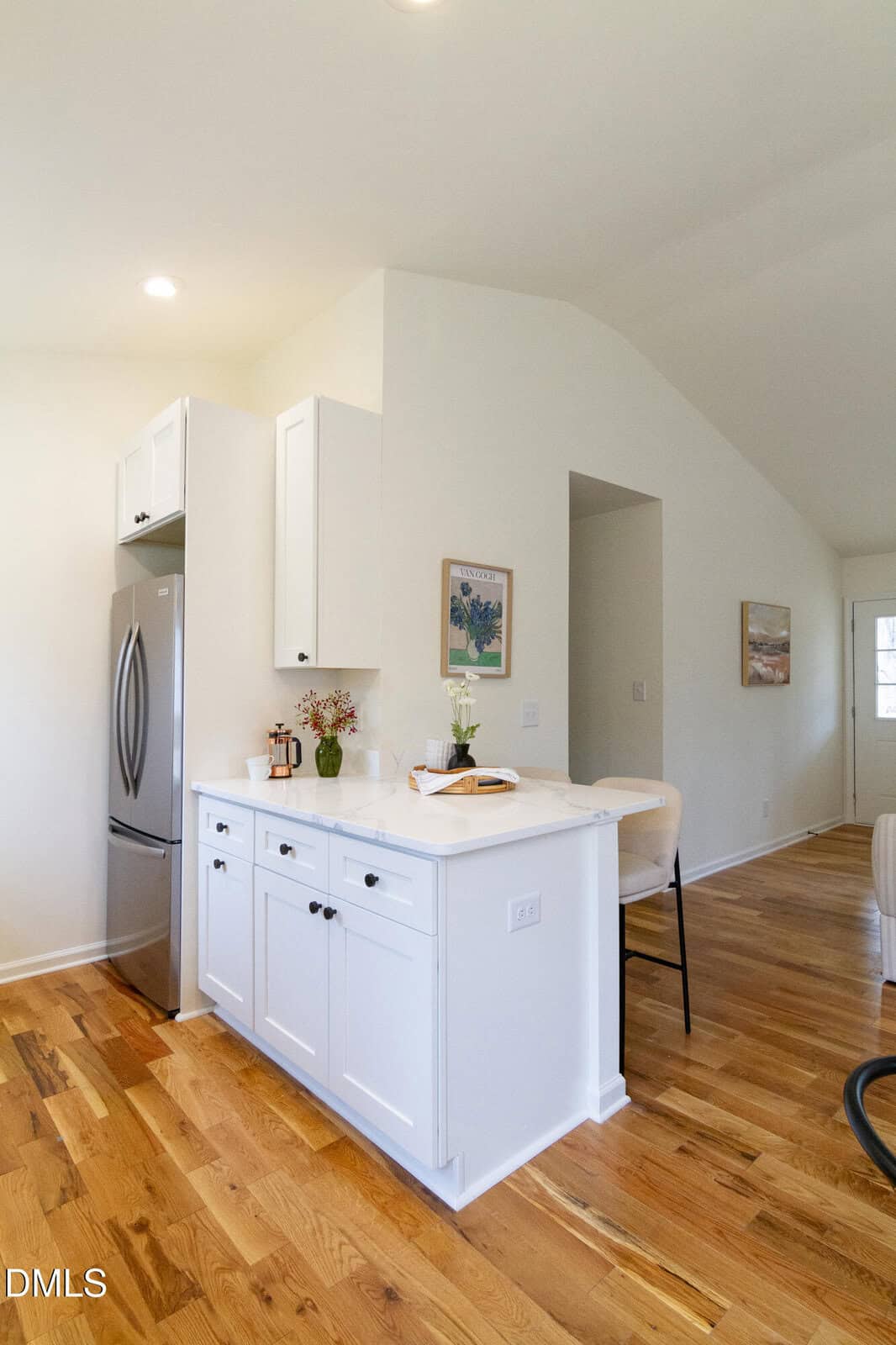 A bright, modern kitchen with white cabinets, stainless steel refrigerator, wooden floors, and simple decor. A countertop with stools and flowers separates the kitchen from the living space. Artwork hangs on light-colored walls.